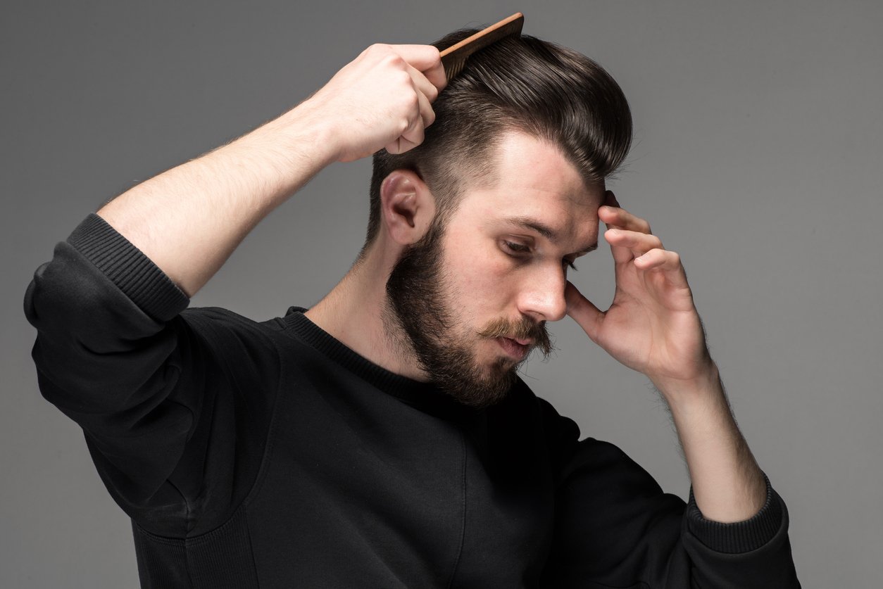 Mann mit braunen Haaren und Bart sieht nach unten und stylt seine Haare mit einem Kamm zurück vor einem grauen Hintergrund